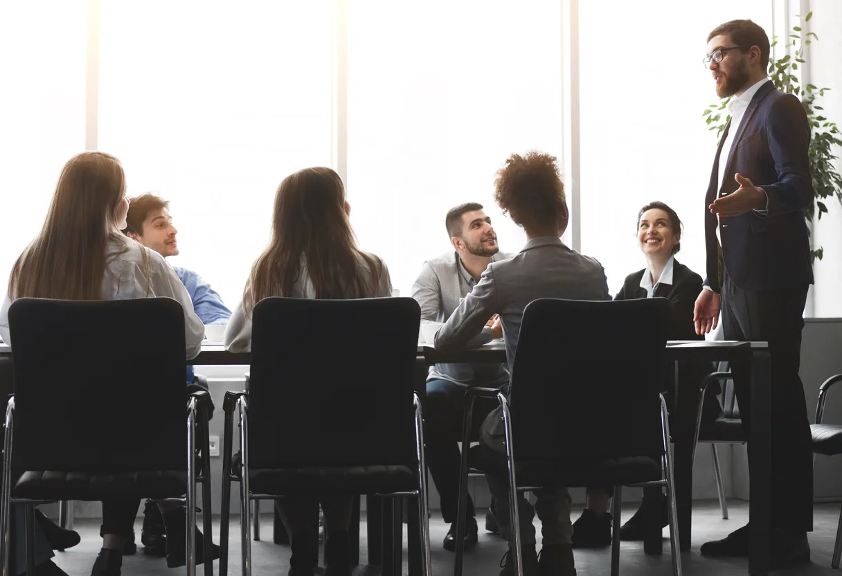 Executive standing to address a seated team of six colleagues at a conference table